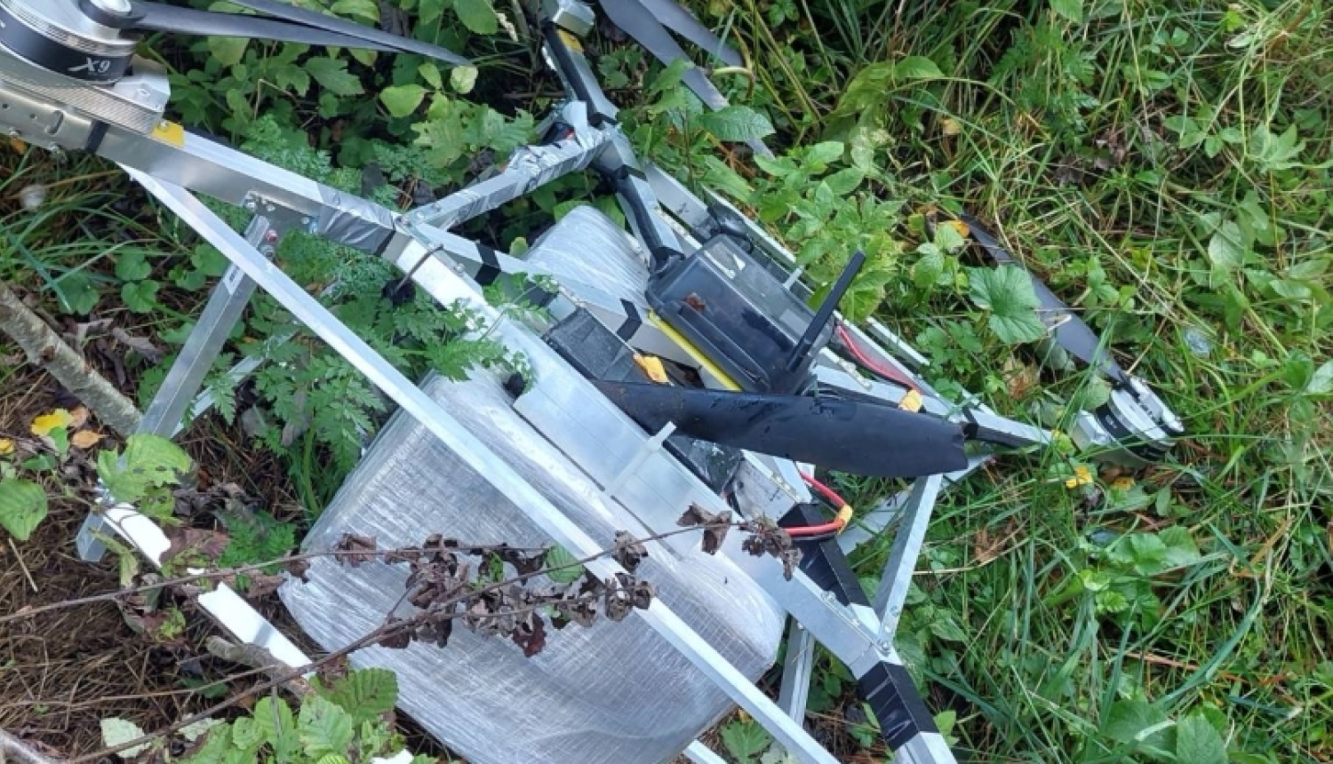 The confiscated drone lying in forest undergrowth in Demene Parish, Latvia, with the white-polyethylene-wrapped cigarette box still strapped to its underside
