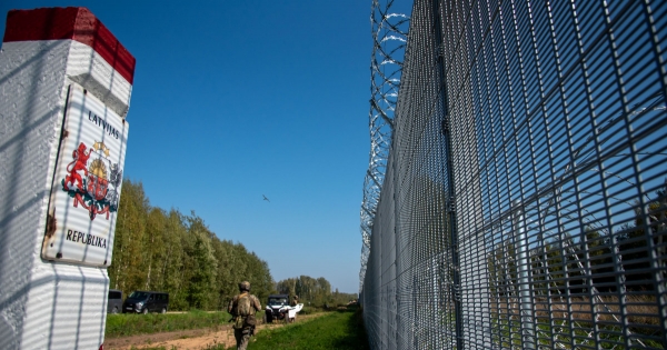 Associative image, Latvian border marker post beside a fence with a patrol vehicle and soldier in the background
